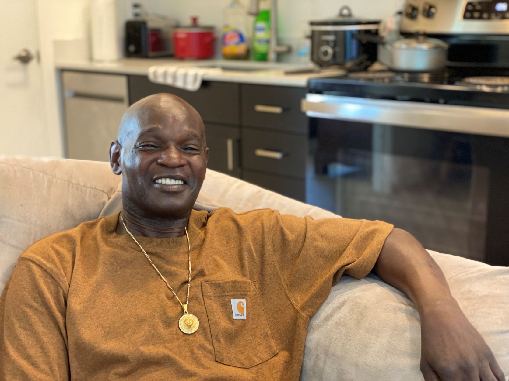 Dale, a Black man with a shaved head wearing a tan t-shirt and a necklace with a round medallion, sits on his sofa smiling.