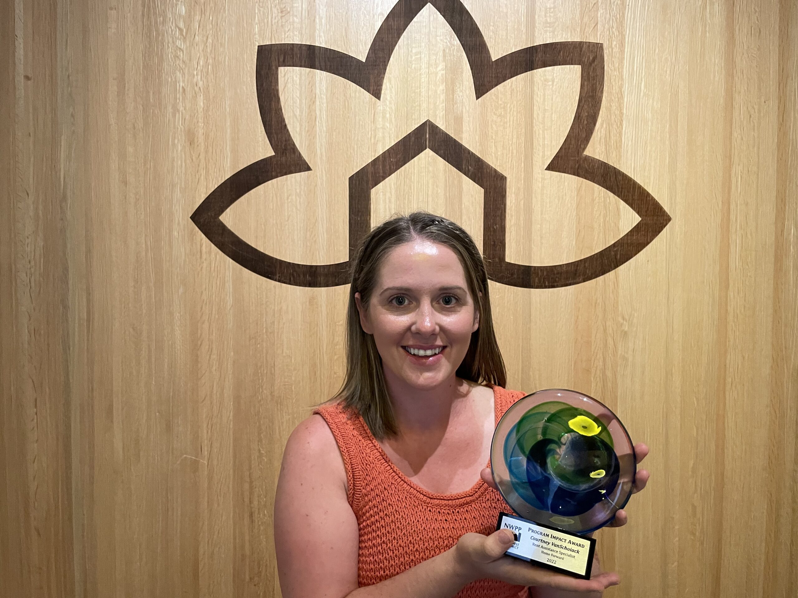Courtney VanSchoaick holds her award, a colored glass disc on a base with a plaque. She stands beneath the Home Forward logo on a wooden wall