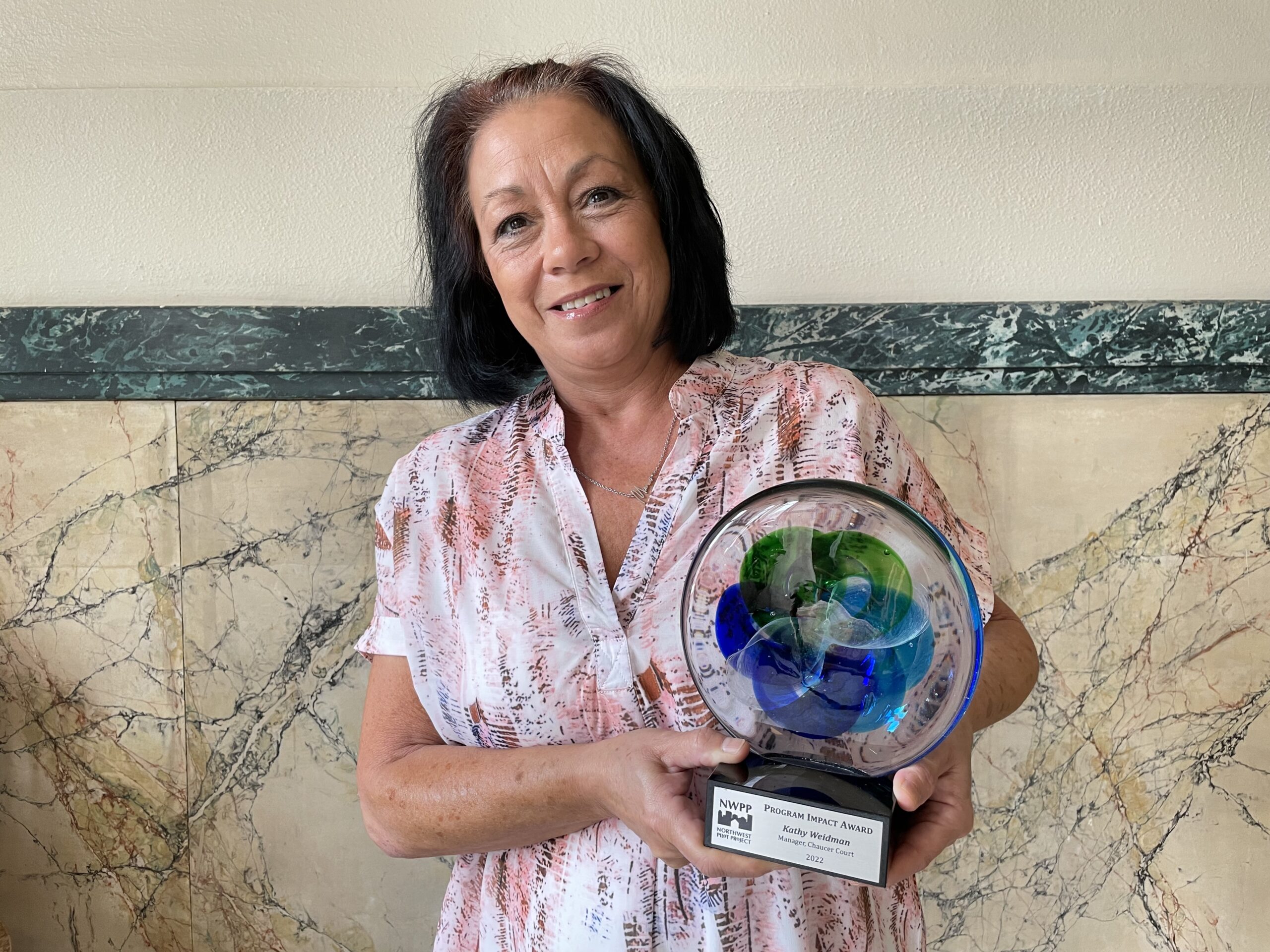 Kathy Weidman holds her award, a colored glass disc on a base with a plaque. She stands in front of a marbled stone wall in the main Chaucer Court stairway