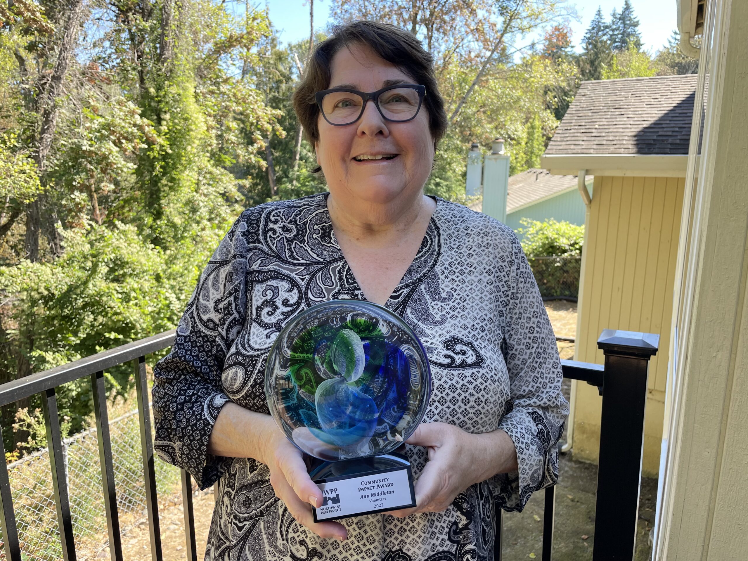 NWPP supporter Ann Middleton holds her 2022 Impact Award. Behind her is a black railing with trees beyond