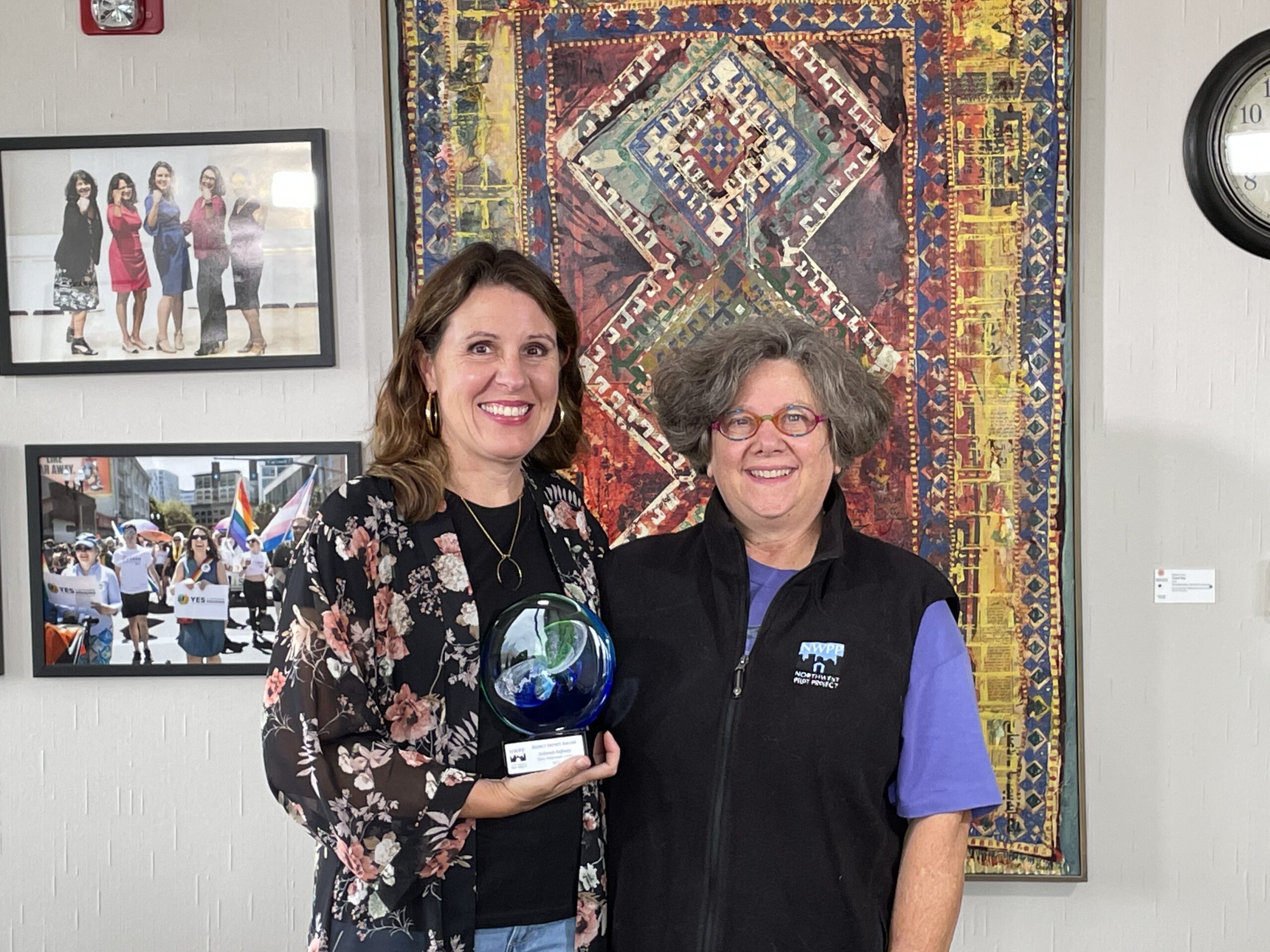 Multnomah County Chair Deborah Kafoury and NWPP executive director Laura Golino de Lovato stand before a colorful tapestry. Chair Kafoury holds the award