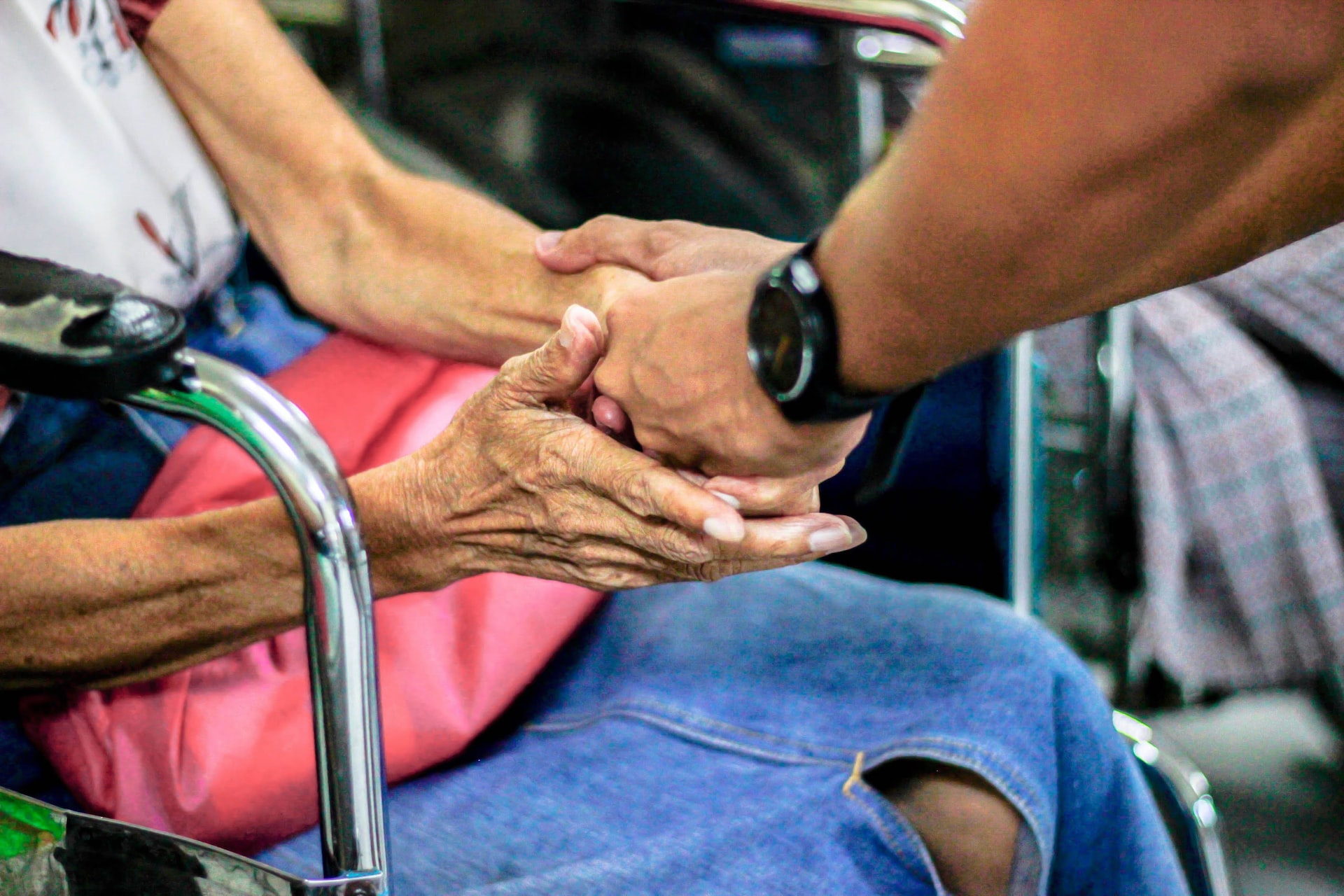 closeup of a seated person, who appears to be an older adult, holding both hands with someone standing in front of them. Only the hands and the lap of the seated person are in the photo.